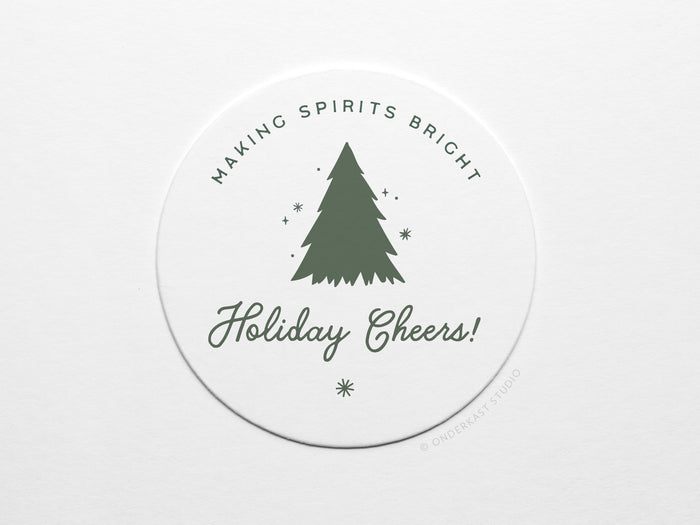 The Holiday Cheers Coaster Set by Onderkast Studio features round white letterpress coasters with a green Christmas tree and stars, “MAKING SPIRITS BRIGHT” above, “Holiday Cheers!” in script below, printed on durable pulp board.