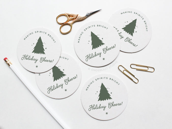 The Holiday Cheers Coaster Set by Onderkast Studio features five round letterpress coasters with a Christmas tree design and “Making Spirits Bright, Holiday Cheers!” text, arranged on a white surface with office supplies to highlight handmade quality.