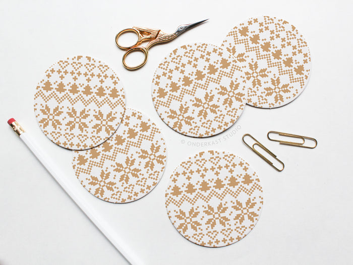 The Holiday Cross Stitch Coaster Set by Onderkast Studio features five round pulp board coasters with a beige snowflake pattern, shown on a white surface with gold scissors, two gold paperclips, and a white pencil with red eraser.