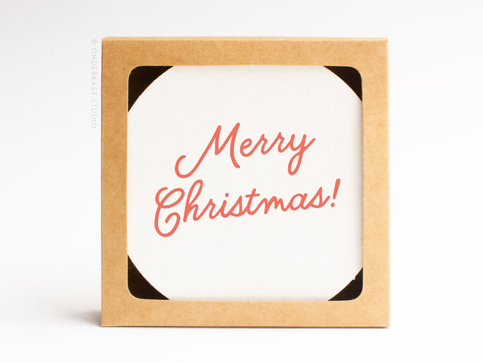 The Merry Christmas Coaster Set by Onderkast Studio comes in a brown, hand-assembled recyclable kraft box with a white card inside that says “Merry Christmas!” in red cursive letters.