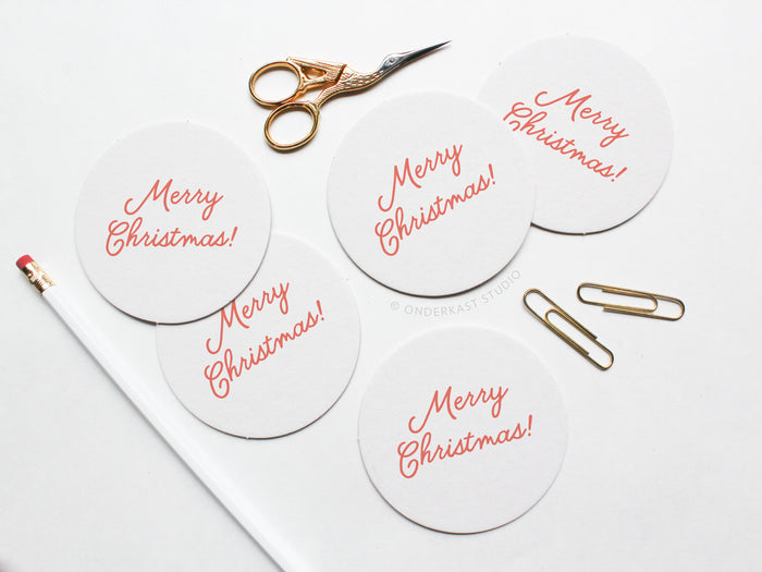 The Merry Christmas Coaster Set by Onderkast Studio features round white letterpress coasters with red text, arranged with gold scissors, a white pencil, and paperclips—ideal for gifting in a recyclable kraft box.