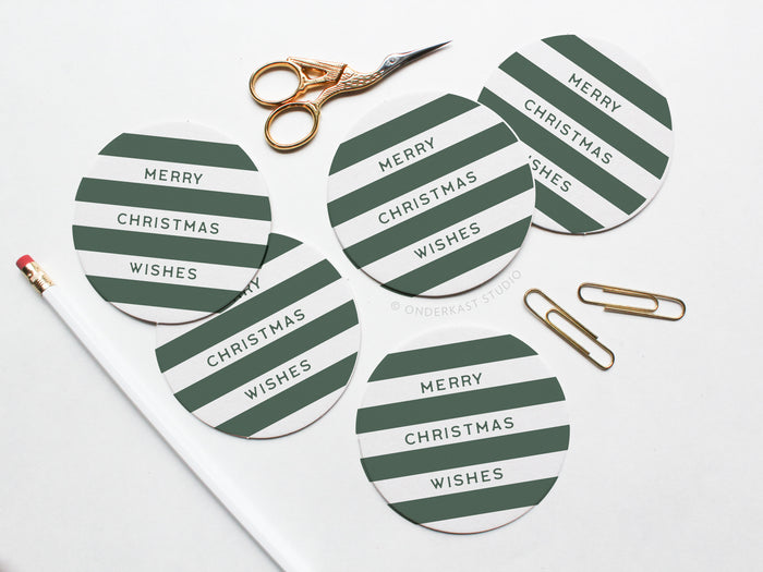 The Merry Christmas Wishes Coaster Set by Onderkast Studio features round pulp board coasters with green and white stripes and “Merry Christmas Wishes” text, displayed on a white surface with a pencil, scissors, and gold paper clips.