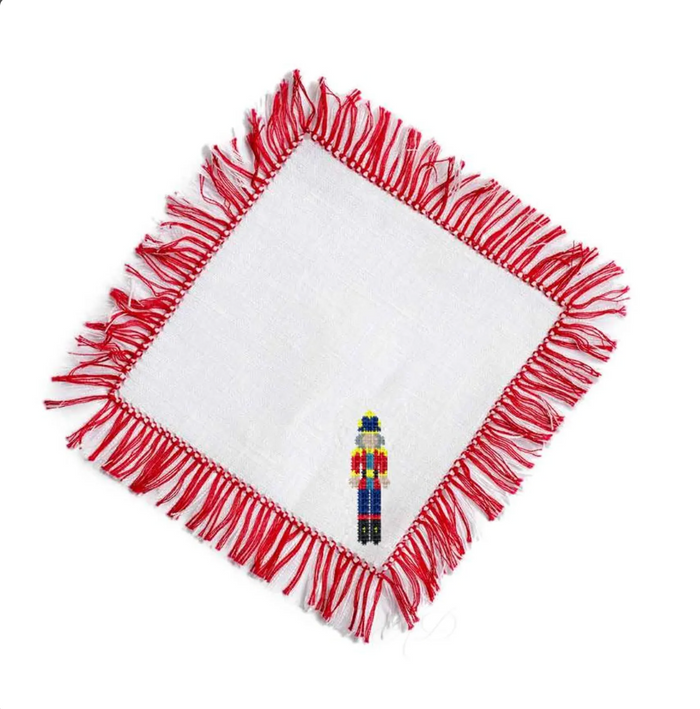 A square white fabric napkin with red fringed edges, shown on a plain white background with a small colorful nutcracker figurine placed near the center.