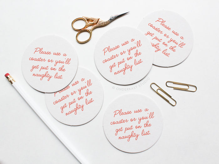 The Naughty List Coaster Set by Onderkast Studio features four round white letterpress coasters with “Please use a coaster or you’ll get put on the naughty list” in red script, made from durable pulp board.