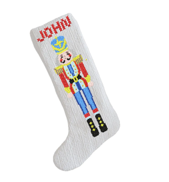 A white knit Christmas stocking featuring a colorful embroidered nutcracker figure and the name 'JOHN' in red at the top, shown on a plain white background.