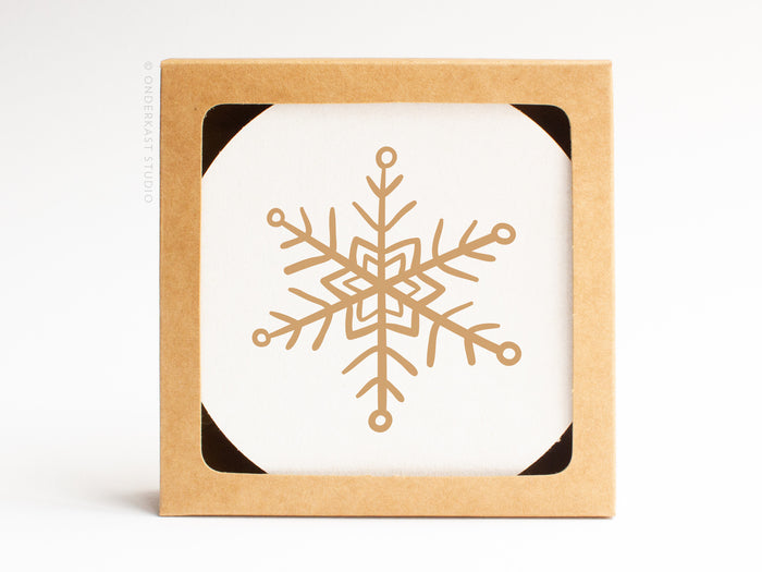 The Snowflake Coaster Set by Onderkast Studio is presented in a square box with a cutout, showcasing a white coaster adorned with a gold geometric snowflake for an elegant, handmade touch.