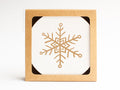 The Snowflake Coaster Set by Onderkast Studio is presented in a square box with a cutout, showcasing a white coaster adorned with a gold geometric snowflake for an elegant, handmade touch.
