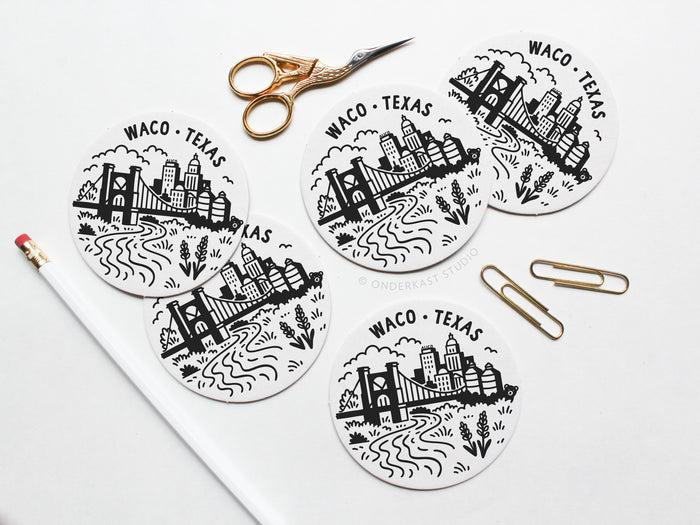 Five round Waco Texas Skyline Letterpress Coasters by Onderkast Studio are arranged on a white surface with gold scissors, a white pencil with a pink eraser, and two gold paper clips nearby.